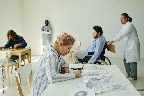 Side view of mature blond female patient of lunatic asylum drawing something on paper sheet while sitting by desk against other people