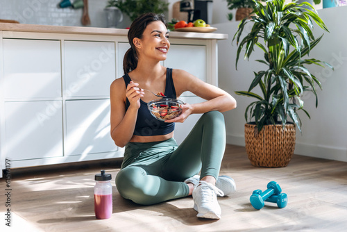 Athletic woman eating a healthy bowl of muesli with fruit sitting on floor in the kitchen at home