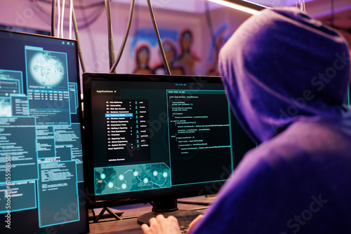Hacker in hood running illegal malware on computer screen with multiple windows. Criminal launching malicious software and coding ransomware in abandoned warehouse at night