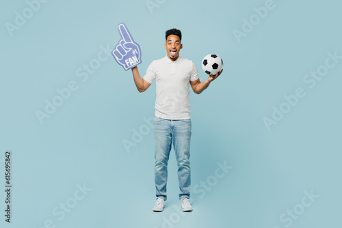 Full body young man fan wear basic t-shirt foam 1 fan glove finger up cheer up support football sport team hold in hand soccer ball watch tv live stream isolated on plain pastel blue color background.