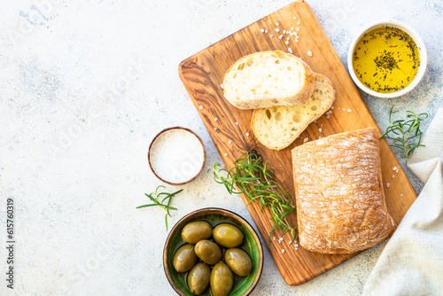Ciabatta bread on wooden board with olive oil, olives and herbs. Top view on white table.