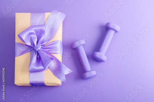 Holiday birthday party sport flat lay composition with purple dumbbells and craft gift with lilac bow on violet paper background. Top view, horizontal orientation