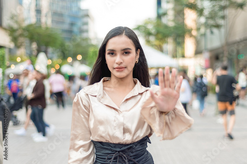 A stern young woman politely says no, gesturing to stop with her open palm. Urban city plaza with crowd setting.