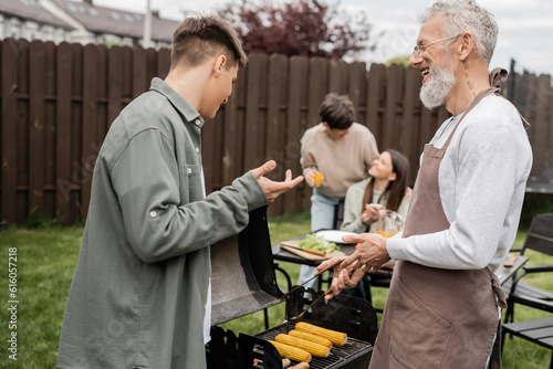 tattooed and bearded father and son preparing food on bbq grill, grilling corn, communication, family bbq party, middle aged, happy parents day, translation of tattoo: harm none do what you will