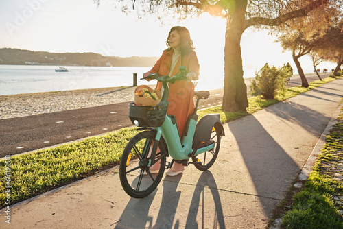 Pregnant woman in orange coat on electric bicycle on the promenade at sunset
