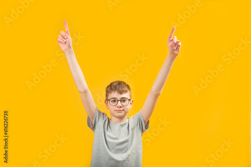 Brunette boy wears glasses and gray T-shirt raised his hands up shows with index fingers up. Isolated on yellow background. Copy space.