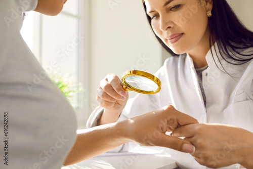 Professional dermatologist examining patient. Doctor holding magnifying glass and investigating growth on hand of young woman. Dermatology, skin health, cancer screening, melanoma prevention concept