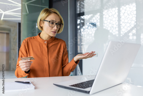 Sad and unhappy woman inside workplace office with laptop holding bank credit card, businesswoman cheated got negative account balance and online money transfer error.