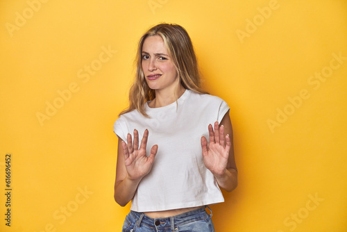 Young blonde Caucasian woman in a white t-shirt on a yellow studio background, rejecting someone showing a gesture of disgust.