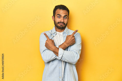 Casual young Latino man against a vibrant yellow studio background, points sideways, is trying to choose between two options.