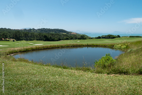 Lago de água a meio de um verde campo de golfe com algumas árvores e o mar ao fundo num dia ensolarado