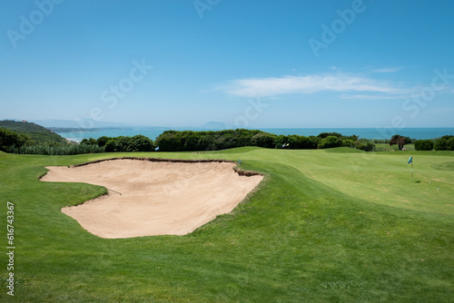 Bunkers ou espaços de areia a meio a um verde campo de golfe com algumas árvores e o mar ao fundo num dia ensolarado