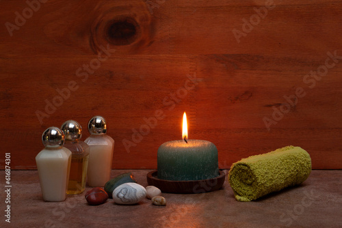 Bath elements: shampoo, cream, green towel, stones and aromatic candle on a wooden background