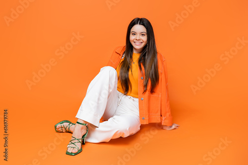 Fashionable and smiling brunette teenage girl in heels and denim jacket looking at camera while sitting and posing on orange background, cool and confident teenage girl, gen z fashion