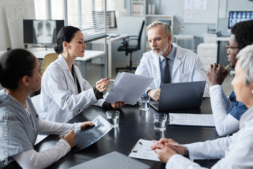 Young female clinician showing medical documents to experienced male doctor sitting by table in front of group of intercultural colleagues