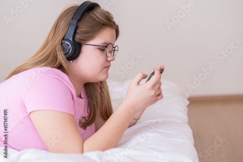 Relaxed teen girl checking messages on smartphone listens to music in headphones overweight teenager lying on bed enjoys social networks and favorite song at home