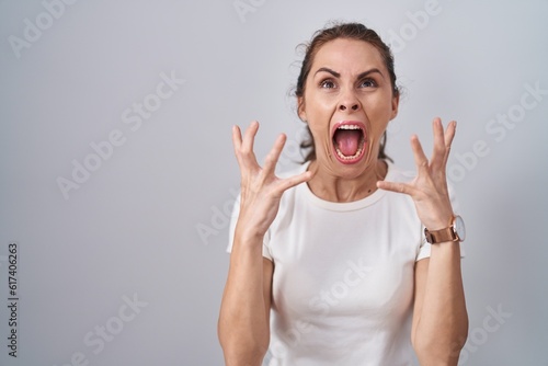 Beautiful brunette woman standing over isolated background crazy and mad shouting and yelling with aggressive expression and arms raised. frustration concept.