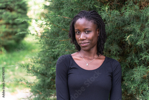 Young black woman with natural sister loc hairstyle smiling while standing in front of a pine tree at a park. No make up and wearing a black top