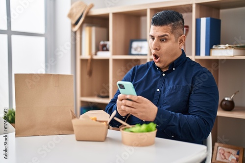 Hispanic young man eating take away food using smartphone in shock face, looking skeptical and sarcastic, surprised with open mouth