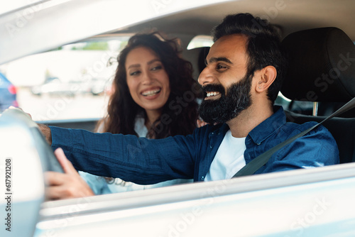 Smiling arab woman looking at male at steering wheel, talking and smiling, enjoying road travel in their new car