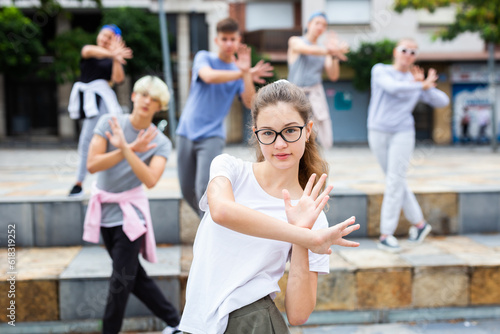 Positive girl dancing modern choreography with group of teenagers on city street on summer day.