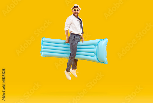 Happy funny man in office clothes, beach sun hat and sunglasses riding on blue inflatable water mattress and flying on yellow studio background. Summer, holiday, vacation, fun concept