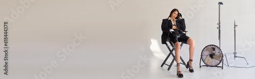 studio photography, young asian woman in blazer, white shirt and latex shorts sitting on folding chair near electric fan on grey background, fashion statement, looking away, full length, banner