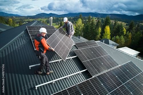 Workers building solar panel system on roof of house. Men technicians in helmets carrying photovoltaic solar module outdoors. Concept of alternative and renewable energy.