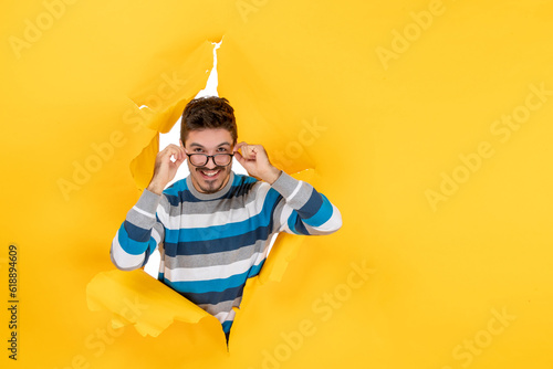 front view young man taking off eyeglasses peeping through hole in paper yellow wall