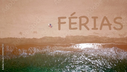 Vista aérea de uma bela praia de areia junto ao mar azul, onde dois veranistas estão deitados e tomando banho de sol, ao lado deles a inscrição "FÉRIAS" na areia