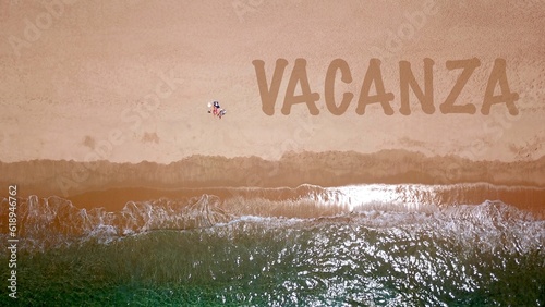 Vista aerea di una bellissima spiaggia sabbiosa in riva al mare blu, dove due vacanzieri sono sdraiati e prendono il sole, accanto a loro la scritta "VACANZA" nella sabbia