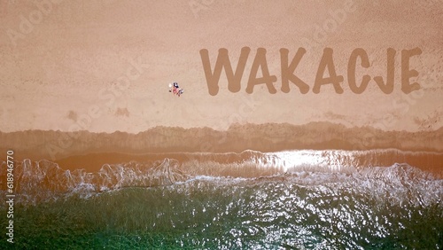 Widok z lotu ptaka na piękną piaszczystą plażę nad błękitnym morzem, gdzie leży i opala się dwóch wczasowiczów, obok nich napis "wakacje" na piasku