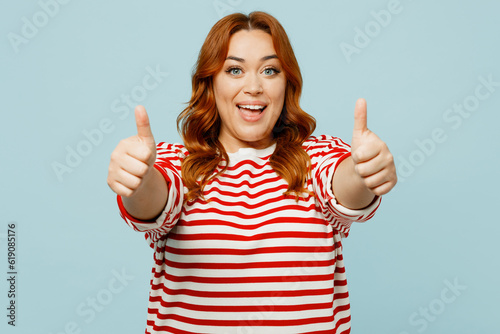 Young happy smiling fun chubby overweight woman wear striped red shirt casual clothes showing thumb up like gesture isolated on plain pastel light blue background studio portrait. Lifestyle concept.