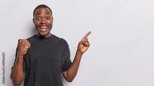 Dark skinned African man clenches his fist in celebration of good news and wonderful offer with beaming expression points index finger towards empty space wears black t shirt isolated over white wall