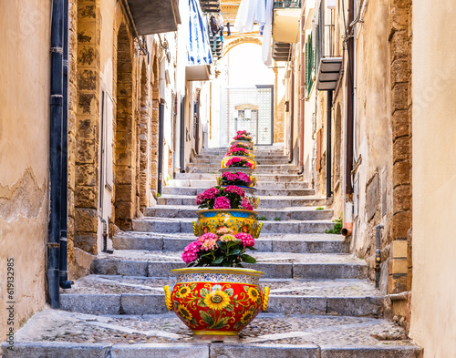 Narrow street in the old town of Cefalu, medieval village on Sicily island, Italy. Flower pots with traditional sicilian decoration. La Rocca cliff. Popular tourist attraction in Province of Palermo
