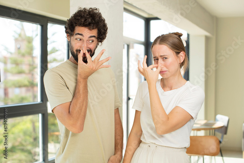 young adult couple feeling disgusted, holding nose to avoid smelling a foul and unpleasant stench