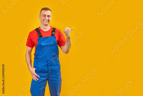 Smiling male foreman engineer wearing blue overalls and points with his finger to the side. Yellow background. Copy space.