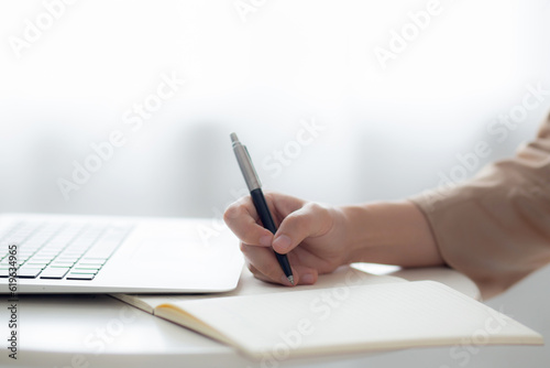 Closeup hands young asian woman writing on notebook for planning working and using laptop computer on desk at home, female notes about finance, female study, business and communication concept.