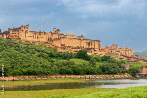 Amer fort palace with beautiful domes, arches, and balconies stands majestically amid a blue cloudy sky on the top of the mountain located near the lake surrounded by green grassland. Rajasthan, India