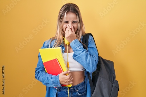 Young blonde woman wearing student backpack and holding books smelling something stinky and disgusting, intolerable smell, holding breath with fingers on nose. bad smell