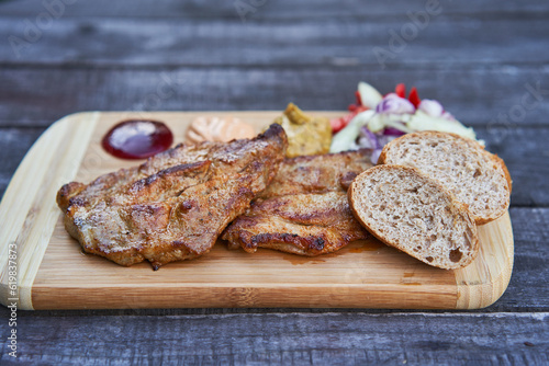Two grilled barbeque pork collar steaks served outside in garden restaurant on the wooden plate with slices of whole grain bread, choped onions, tomatoes, mustard and barbeque sauces. Tasty party food