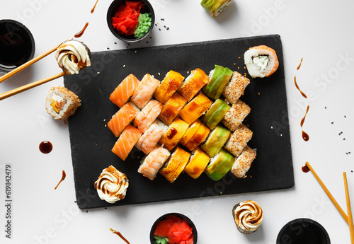 Flat lay of various sushi rolls placed on stone board with chopsticks and soy sauce on gray background
