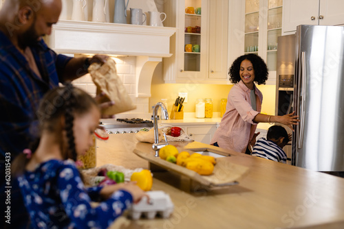 Happy biracial parents, son and daughter unpacking grocery shopping in kitchen and stocking fridge