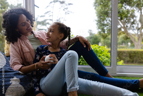 Happy biracial mother and daughter sitting by window to sunny garden and talking at home, copy space