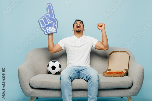 Full body winner young man fan wear basic t-shirt foam 1 fan glove finger up cheer up support football sport team holding soccer ball sit on sofa watch tv live stream isolated on plain blue background