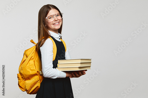 Side view of excellent schoolgirl holding stack of books in hands, wearing glasses and yellow backpack, isolated over white background with copy space