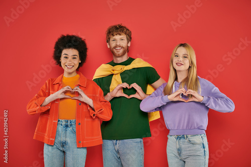 open relationship, polygamy concept, three interracial lovers showing heart sign with hands on coral background, cultural diversity, polyamorous, happy multiethnic people looking at camera