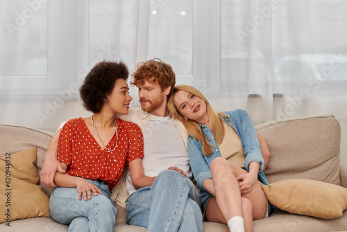 modern family, polygamy concept, freedom in relationship, cultural diversity, redhead man sitting with multicultural women on couch in living room, polyamorous lifestyle, non traditional