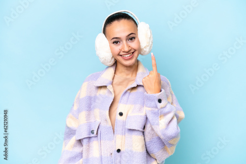 Young Arab woman wearing winter muffs isolated on blue background pointing with the index finger a great idea