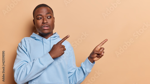 Photo of serious dark skinned adult man with beard points fingers aside on copy space dressed in casual blue sweatshirt gives recommendation isolated over brown background. Advertisement concept